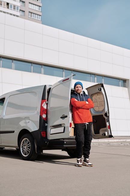 A man wearing a blue beanie, red and navy jacket, and black trousers stands with arms crossed outside a light grey delivery van parked on a paved area in front of a modern, multi-storey building with curved white panels and narrow windows. The van's side and rear doors are open, revealing an empty cargo space with visible cardboard boxes and packing materials inside. The scene suggests a home relocation or furniture transport process, with the man representing a worker from Man with Van Ilford, part of their removals services. The lighting indicates daytime with natural sunlight, and the setting emphasizes professional loading or unloading activities related to house removals, packing, and moving logistics.