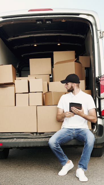 A man wearing a blue beanie, red and navy jacket, and black trousers stands with arms crossed outside a light grey delivery van parked on a paved area in front of a modern, multi-storey building with curved white panels and narrow windows. The van's side and rear doors are open, revealing an empty cargo space with visible cardboard boxes and packing materials inside. The scene suggests a home relocation or furniture transport process, with the man representing a worker from Man with Van Ilford, part of their removals services. The lighting indicates daytime with natural sunlight, and the setting emphasizes professional loading or unloading activities related to house removals, packing, and moving logistics.