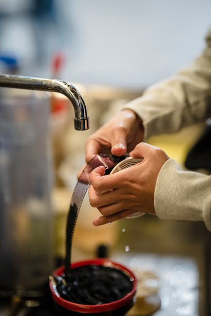 Close-up of a person’s hands filling a clear measuring tape with a silver metallic end, while standing beside a stainless steel sink with a curved faucet in an indoor setting. The person is wearing a beige long-sleeve garment, and the background shows blurred kitchen or workshop elements. This image highlights the careful handling and measurement process involved in home relocation or furniture transport during packing and moving activities. Man with Van Ilford’s removal team uses precise measuring tools to assist with the safe and efficient loading process for interior furniture removal on Cranbrook Road, Ilford, where narrow access can present logistical challenges for moving large items or appliances. The scene emphasizes attention to detail in planning and executing successful house removals, ensuring secure packing and transport of belongings.