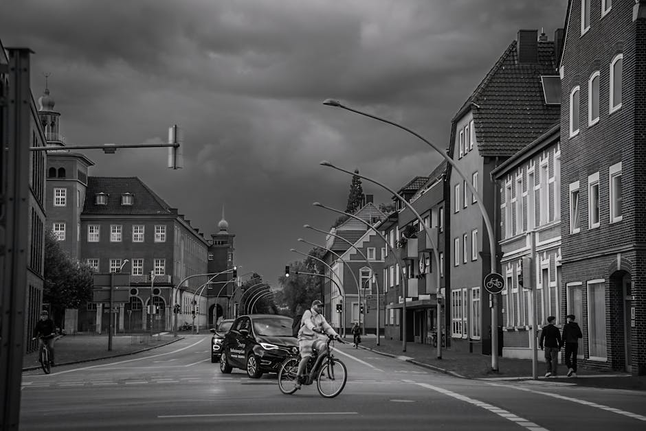 A black and white photograph of a residential street scene in Ilford during daytime, showing the loading process for a house relocation. In the foreground, a person is riding a bicycle across the crosswalk, wearing casual clothing and a helmet. Behind them, a dark vehicle is mounted with a moving company's logo, possibly Man with Van Ilford, loaded with cardboard moving boxes, some wrapped in plastic or fabric for protection. To the right, two pedestrians are walking along the pavement near a bicycle lane sign, while to the left, another cyclist is riding along the street. The street is lined with multi-storey buildings constructed from brick and other materials, featuring large windows, pitched roofs, and decorative architectural elements. Overhead, curved streetlights and several street signs are visible, and in the background, a traffic light hangs above the intersection, with dark clouds overhead suggesting an approaching storm. The scene reflects the logistics involved in furniture transport and packing during an office or home relocation, illustrating the typical street environment during moving day with vehicles, packing materials, and active pedestrians, aligned with the house removals services provided by Man with Van Ilford.