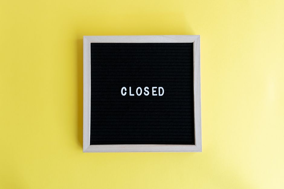 A black felt letter board with a white wooden frame, mounted on a bright yellow wall, displaying the word 'CLOSED' in white plastic letters. The board appears to be used for signage, possibly in a retail or office setting, indicating that the premises are not open for access. This image may relate to home or office closures during a moving process or service restrictions, as handled by Man with Van Ilford, a professional removals company. The yellow background provides high contrast, ensuring the sign's message is clear and easily visible. The clean, minimal design emphasizes the closure notification, with no other objects or furnishings present in the image, highlighting the focus on the sign itself and its communication role within a relocation or closure context.