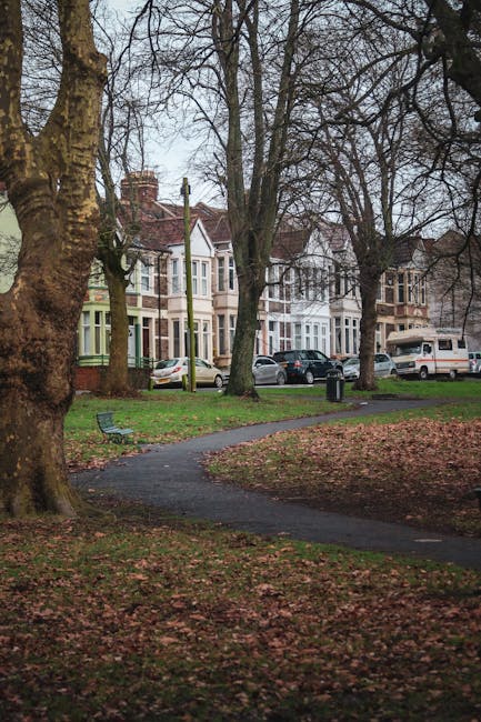 The image shows a residential street within Valentines Park in Ilford, featuring a paved pathway winding through a grassy area with fallen autumn leaves. In the background, there are traditional mid-19th to early 20th-century terraced houses with bay windows, painted in shades of white, cream, and pastel colours, some with front gardens and parked cars lining the street. Tall, leafless trees with thick trunks border the pathway, providing a natural canopy and a sense of quiet in the neighbourhood. A black trash bin is positioned along the pavement, and the scene is illuminated by natural daylight under an overcast sky, suggesting a calm day suitable for home relocation activities. Occasionally, Man with Van Ilford utilises such locations as part of their house removals and furniture transport services, ensuring a smooth packing and loading process for local moves, with moving trucks likely parked nearby for efficient loading of furniture and boxes wrapped in plastic and cardboard for protection during transit.
