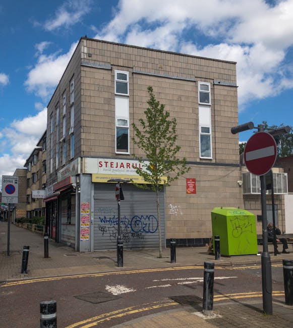 The exterior of a multi-story building with a beige brick facade, located on a street corner. The ground floor features a closed shop with a grey metal shutter covered in graffiti, and a sign above displaying the name 'Stejarl'. A small, young tree is planted at the sidewalk near the corner, partially obscuring the shop front. To the right, there is a bright green public bin with graffiti, a black street sign, and a red and white no-entry traffic sign. The pavement is lined with black bollards, and a bench is visible further to the right. The sky above is partly cloudy with patches of blue, and the scene is captured in natural daylight. The image context relates to urban moving processes, such as home relocation or furniture transport, consistent with services provided by Man with Van Ilford, especially when handling packing and loading tasks near Ilford Station.