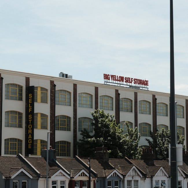 Exterior view of a large multi-storey building with a sign on the roof reading 'Big Yellow Self Storage'. The building has a white facade with vertical brown brick accents and numerous yellow-framed windows arranged in rows. In the foreground, there are several small residential houses with pitched roofs, chimneys, and visible windows, all situated below a streetlamp. Some trees partially obscure the lower part of the building, and the sky above is partly cloudy with a mix of blue and white clouds. The scene illustrates a commercial self-storage facility adjacent to residential properties, captured in natural daylight, relevant to house removals or furniture transport services.