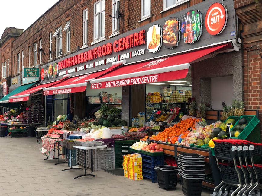Exterior view of a bustling street with a brick building housing the South Harrow Food Centre. The storefront features a prominent red and white sign displaying the shop name, with smaller signs indic