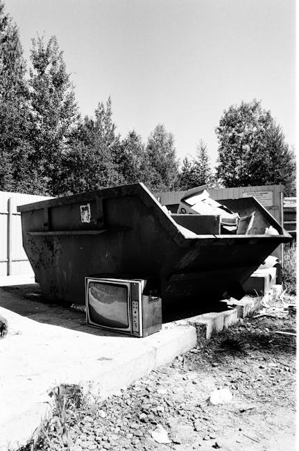 A large, open-top metal dumpster filled with various discarded items including a boxed television, cardboard packaging, and other miscellaneous waste materials, situated on a concrete surface outside