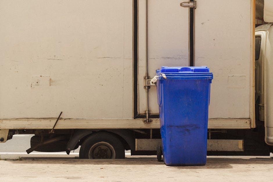 A large, open-top metal dumpster filled with various discarded items including a boxed television, cardboard packaging, and other miscellaneous waste materials, situated on a concrete surface outside